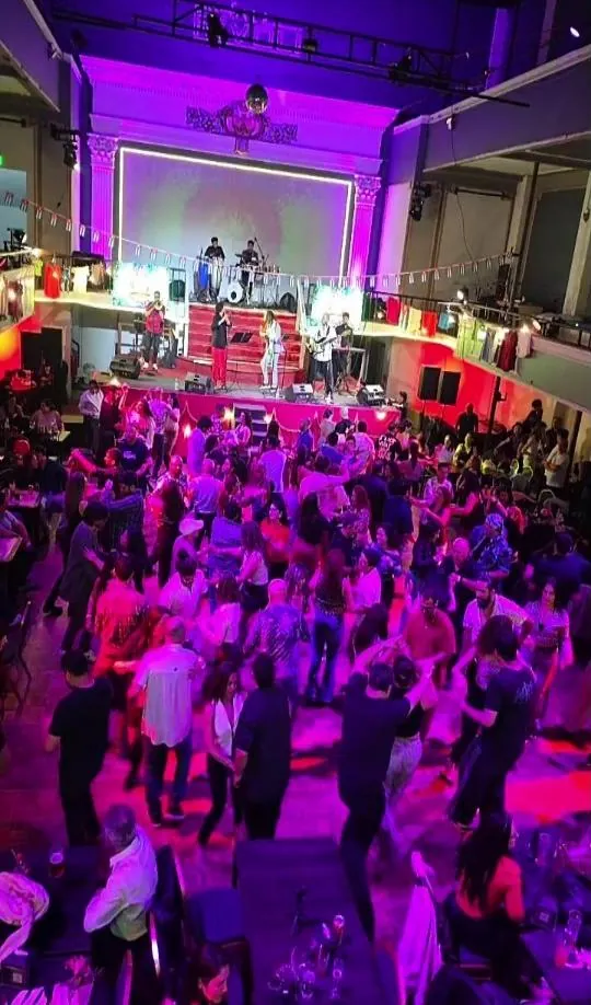 Pista llena de gente bailando frente a una orquesta en vivo, con luces moradas
